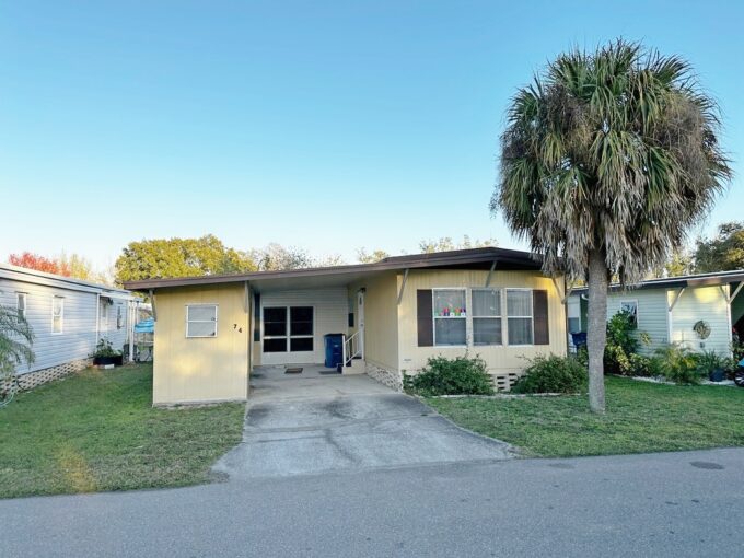 Mellow and Delightful home at 74 Hide A Way LN, Winter Haven, Florida. Yellow vinyl siding with brown shutters, driveway and carport at front entrance, large palm tree in front yard