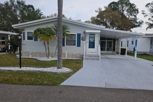 Double-Wide Jacobsen home at 2324 Thoreau DR, Lake Wales, FL where you can enjoy an absolutely joyful retirement lifestyle. Gray Vinyl siding with Blue accent trim on doors and shutters. Gray painted driveway. Clean and crisply landscaped with tropical plants and concrete curbing.