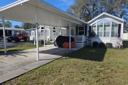 Mobile Comfort: Cozy Retreat for Seniors at 5601 Cypress Gardens RD, Lot M14B in Winter Haven, FL. Single-wide Park Model with side addition. White with covered porch and long carport over driveway.