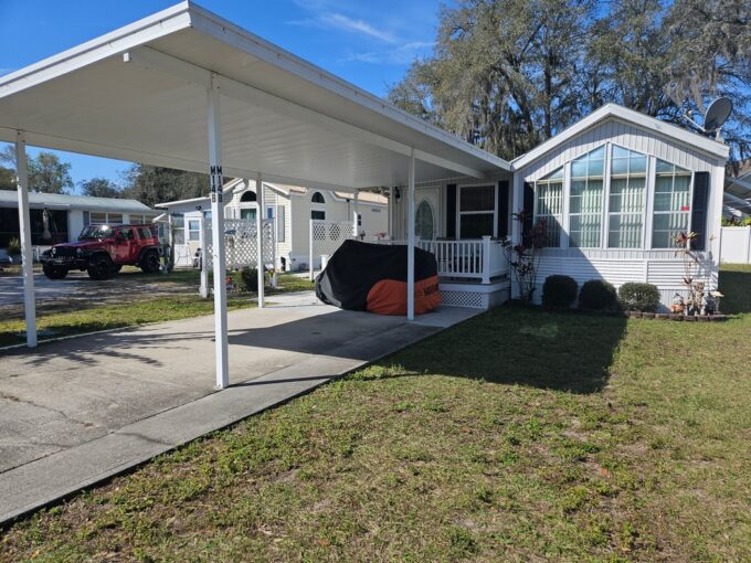 Mobile Comfort: Cozy Retreat for Seniors at 5601 Cypress Gardens RD, Lot M14B in Winter Haven, FL. Single-wide Park Model with side addition. White with covered porch and long carport over driveway.