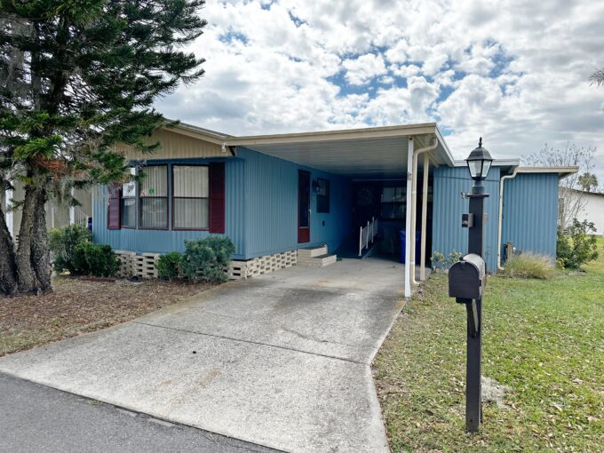 Your Alluring Abode Awaits at 697 Century LN, Winter Haven, FL. Blue single-wide mobile home with ramp to one entrance in carport area off driveway