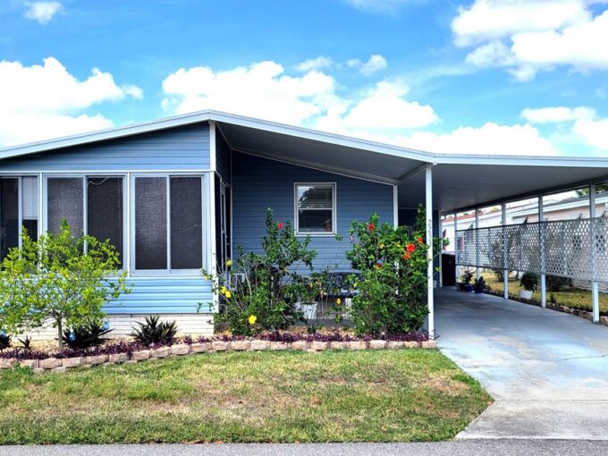 Charming Home featuring an upgraded kitchen and Nestled in Enchanting Community at 2233 Parrot Place, Lake Wales, Florida. Double-Wide manufactured home on with blue siding and white trim, front-facing lanai, landscaped yard and entrance, driveway to carport