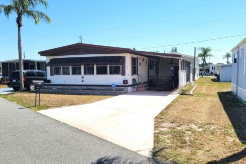 View 2 - exterior picture showing white home with brown accents, front awning to help shade the bank of windows, carport, gutters, private driveway, curbside mail delivery.