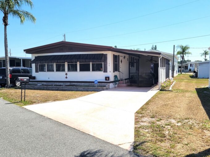 View 2 - exterior picture showing white home with brown accents, front awning to help shade the bank of windows, carport, gutters, private driveway, curbside mail delivery.