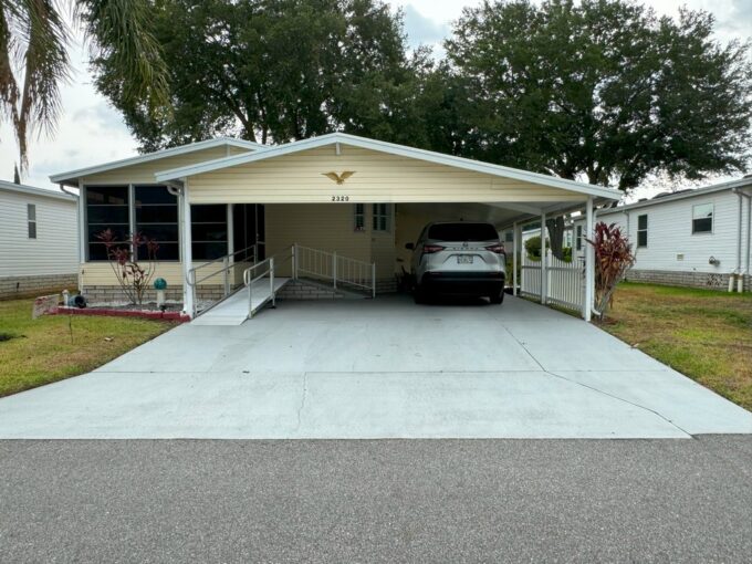 2320 Prestwick Pl, Lot 297 in Winter Haven, FL. Yellow vinyl siding, double driveway with extended carport, ramp, gutters. Mature trees around to offer shade.