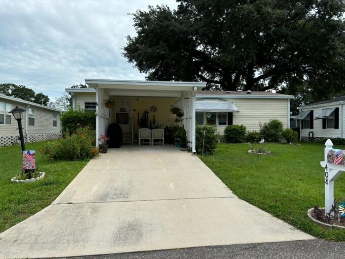 408 Gulf Stream in Kings Pointe, Lake Alfred. Exterior of this duble wide is yellow with white trim. Includes carport, private driveway, curb side mail delivery, gutters, awning and mature landscaping.