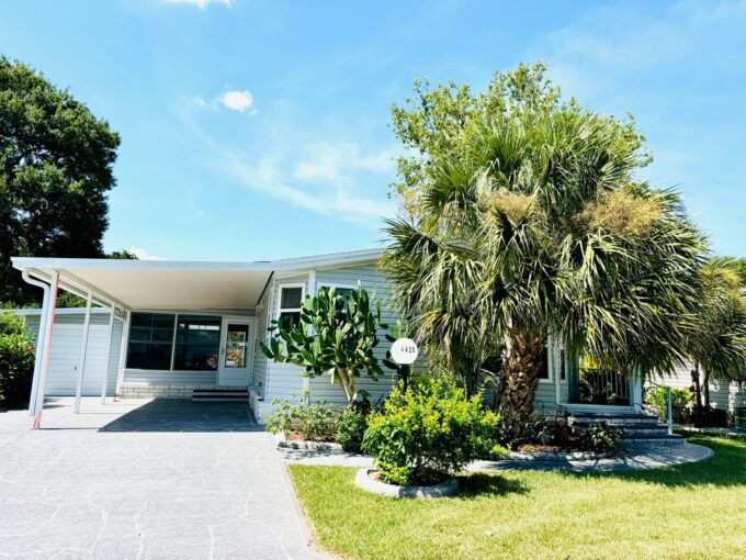 View 2 - Exterior picture shows a small porch at the entry. Oversized private driveway, glass windowed lanai, golf cart garage, carport. Mature landscaping and the home has gutters. Home has Gray siding with white trim.