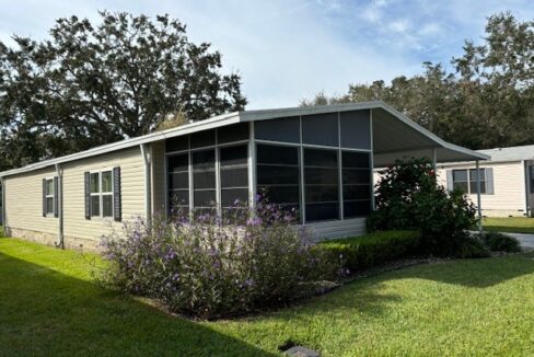 Tan vinyl siding with white trim, front lanai, private driveway, carport with gutters, storage shed at the end of the driveway. Manicured lawn.